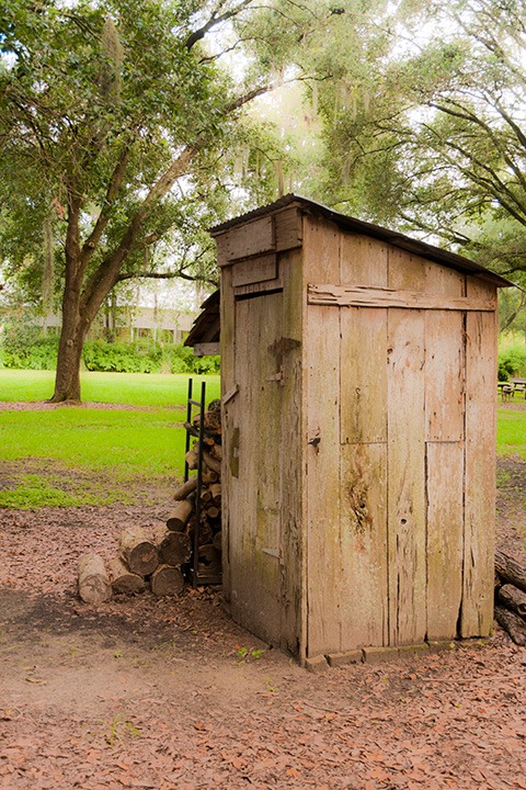 Destrehan Plantation outhouse stands silent beside a woodpile at the edge of a tree-shadowed green
