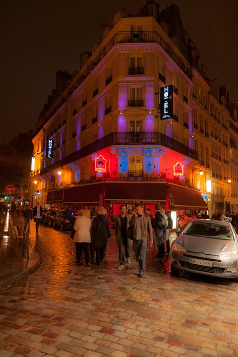 neon-lit Paris flatiron building serves as backdrop for young men walking across glistening cobblestones after a rain