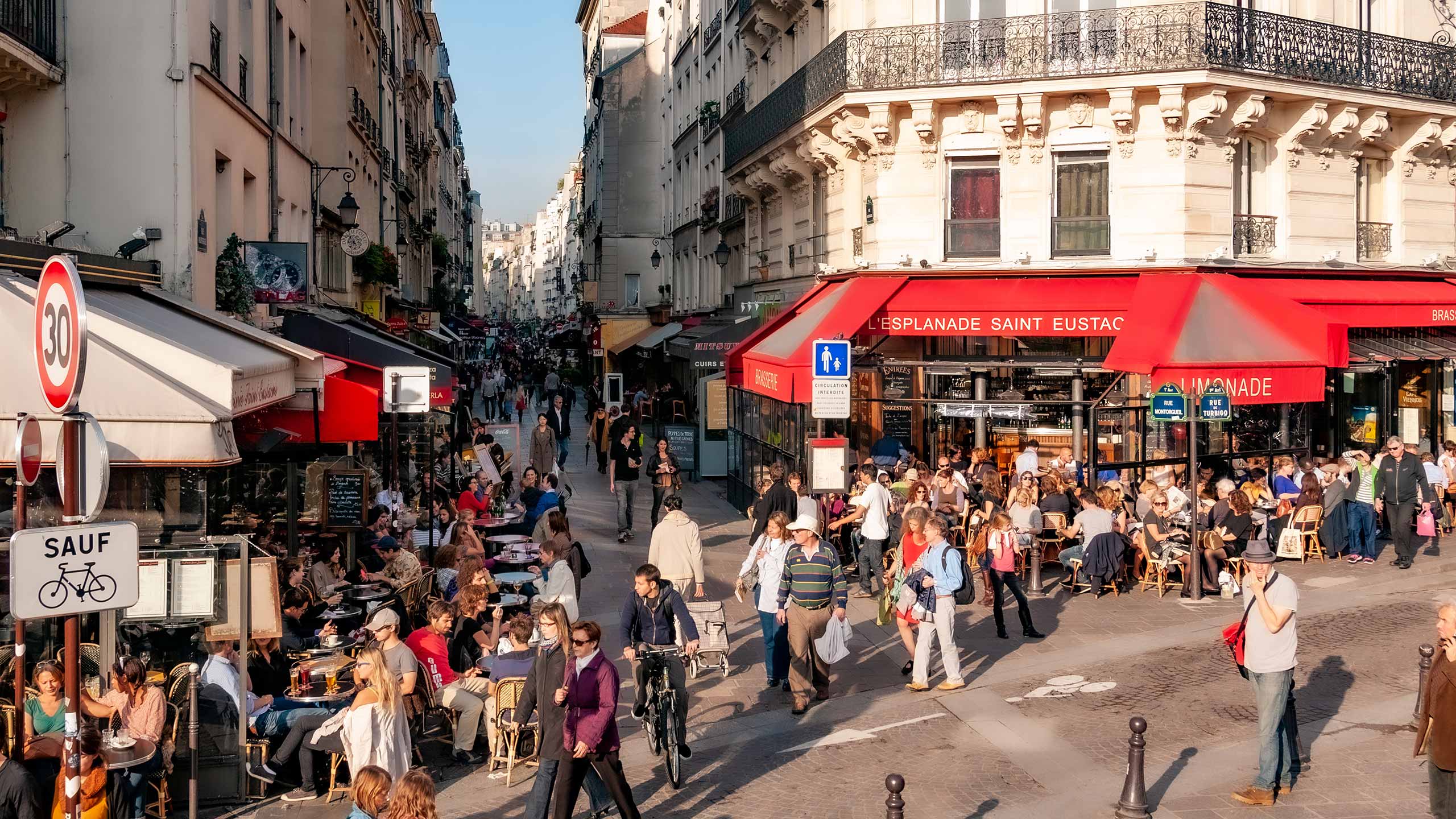 Rue de Turbigo in Paris flanked by stone buildings and filled with people walking, cycling, and sitting at sidewalk cafes as far as the eye can see