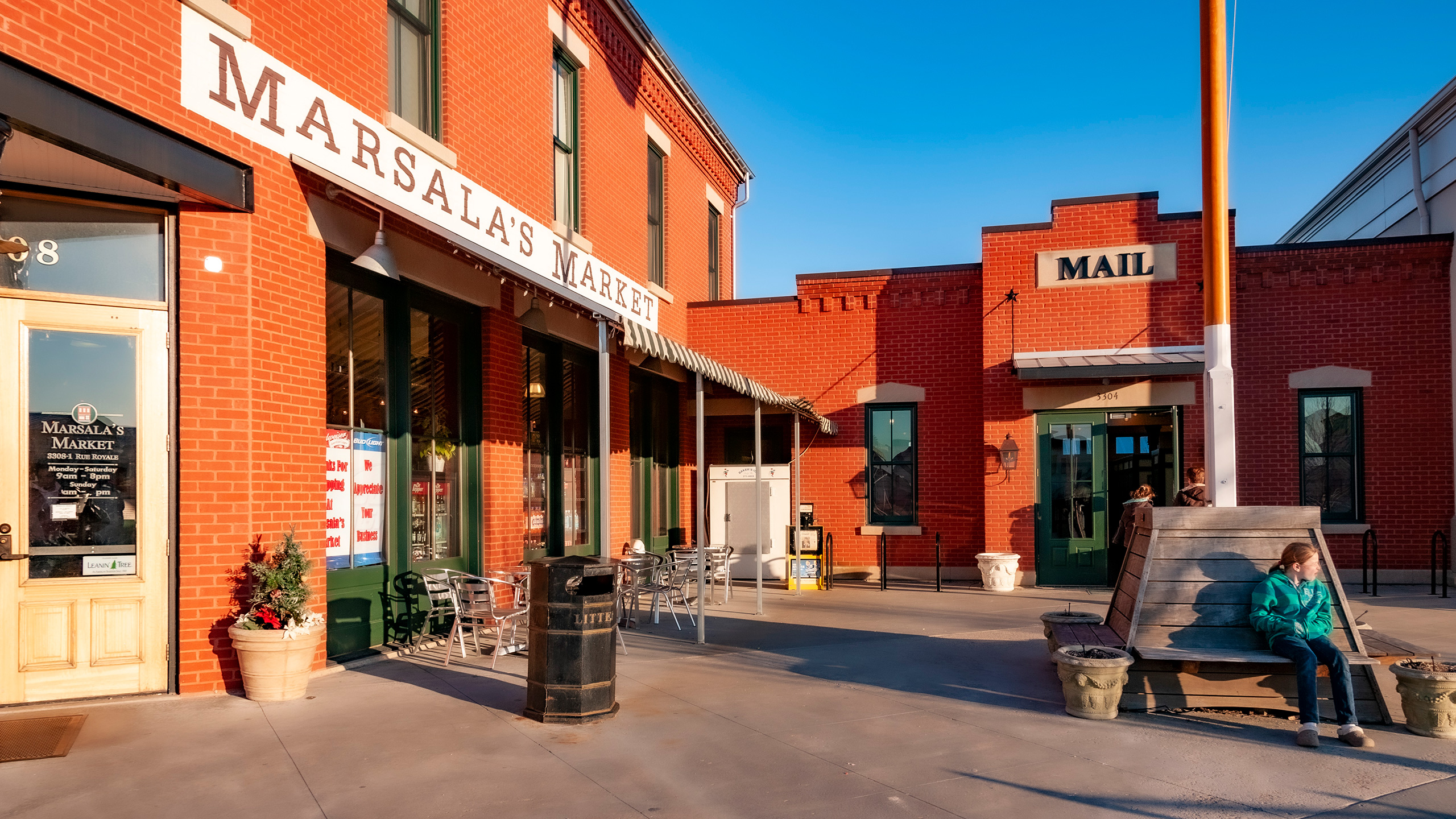 girl sitting beneath flagpole at neighborhood center flanked by market and post office